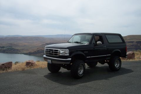 1995 Ford Bronco - A lifted black Ford Bronco sits on a scenic overlook with a lake and hills in the background.
