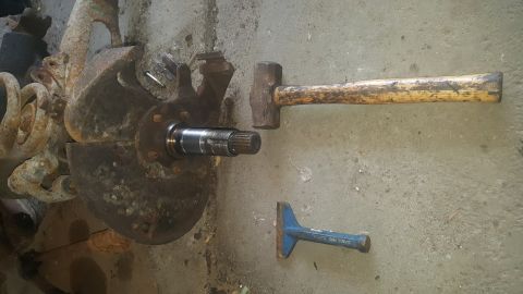 1995 Ford Bronco - A rusty brake rotor and hub assembly is shown on a concrete surface next to a sledgehammer and a blue chisel. A splined shaft protrudes fro…