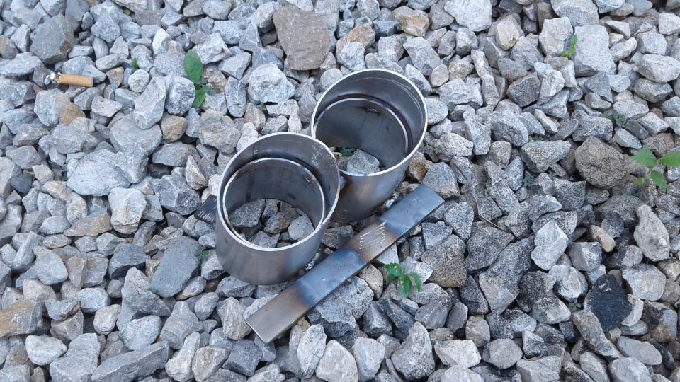 Two metal exhaust pipe adapters and a metal strip on grey gravel.