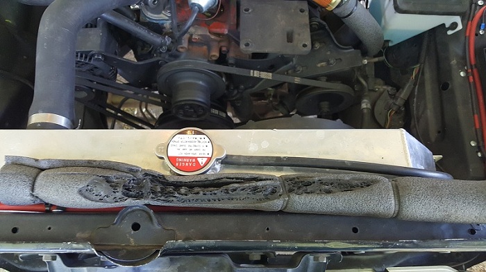 Damaged foam insulation in front of a vehicle radiator and engine with a warning label on the radiator cap.