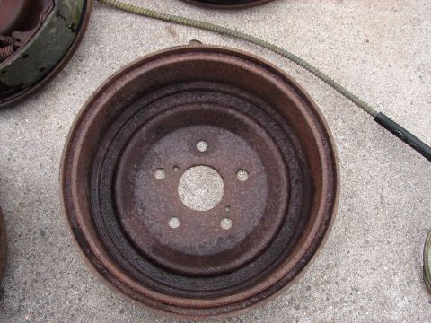 1973 Ford Ranchero - Two rusty brake drums and a brake cable are laid out on a concrete surface. One brake drum is partially visible on the left, showing brake…