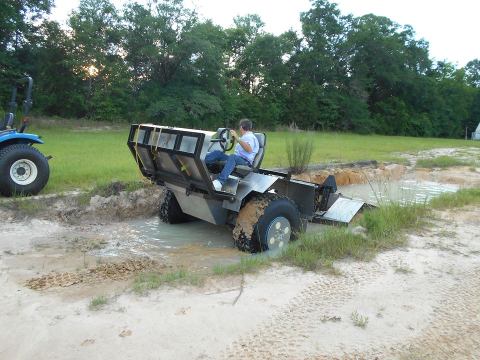 Person driving a custom amphibious vehicle through mud and water.