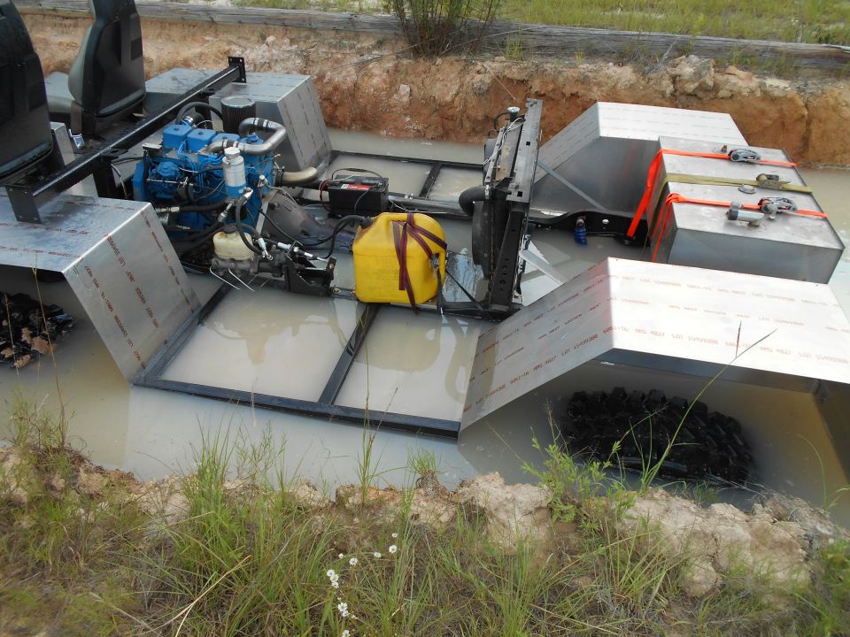 Custom amphibious vehicle with blue engine, metal frame, and pontoons in muddy water.