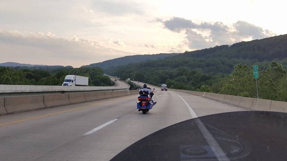 Blue and black cruiser motorcycle on a highway with a wooded hillside.