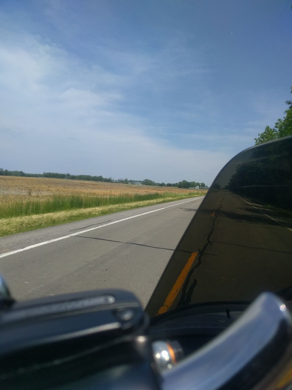 Motorcycle windshield view of a road with a grassy field and trees.