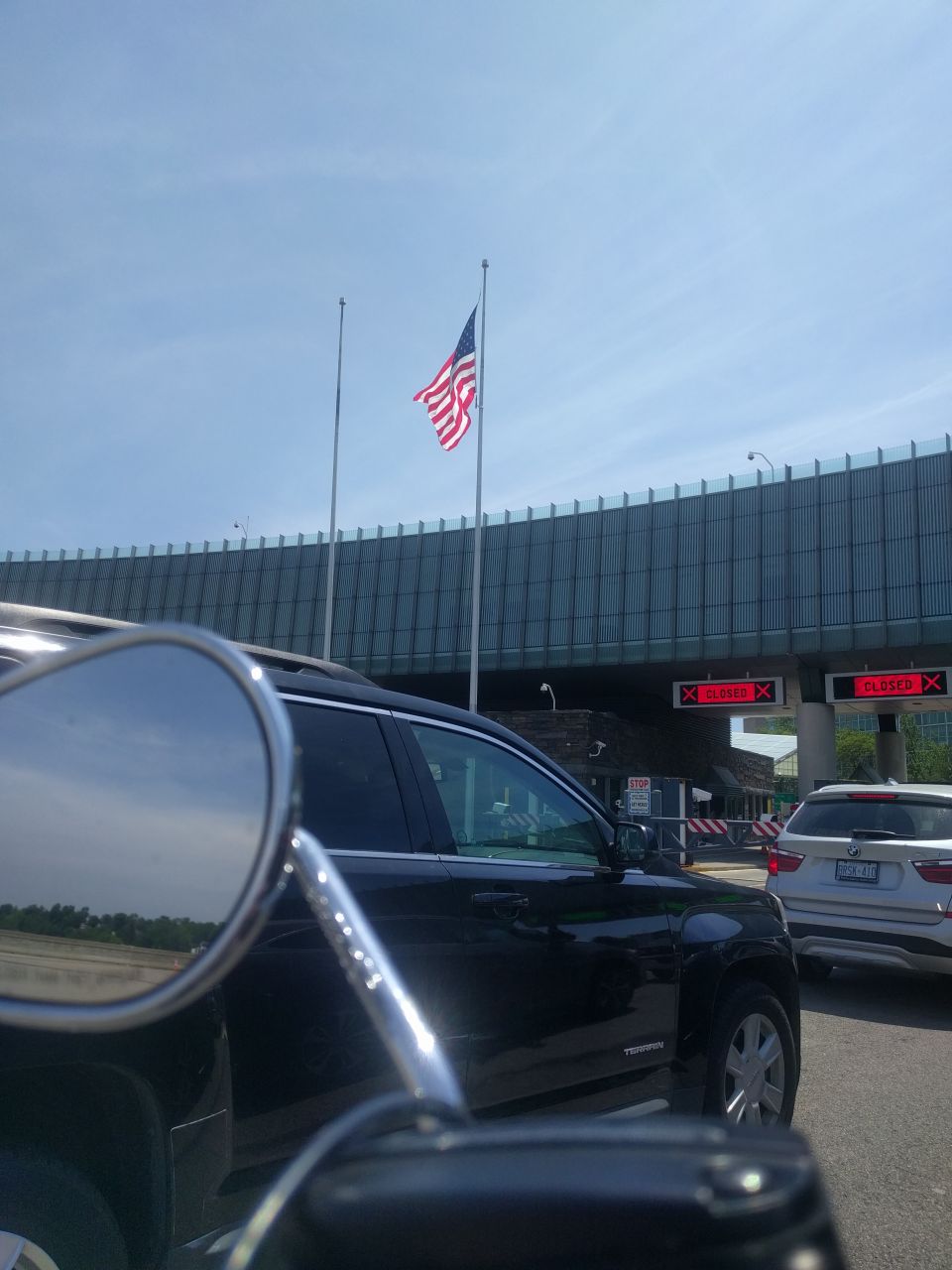 Motorcycle mirror reflecting an SUV and a US flag at a closed border crossing.