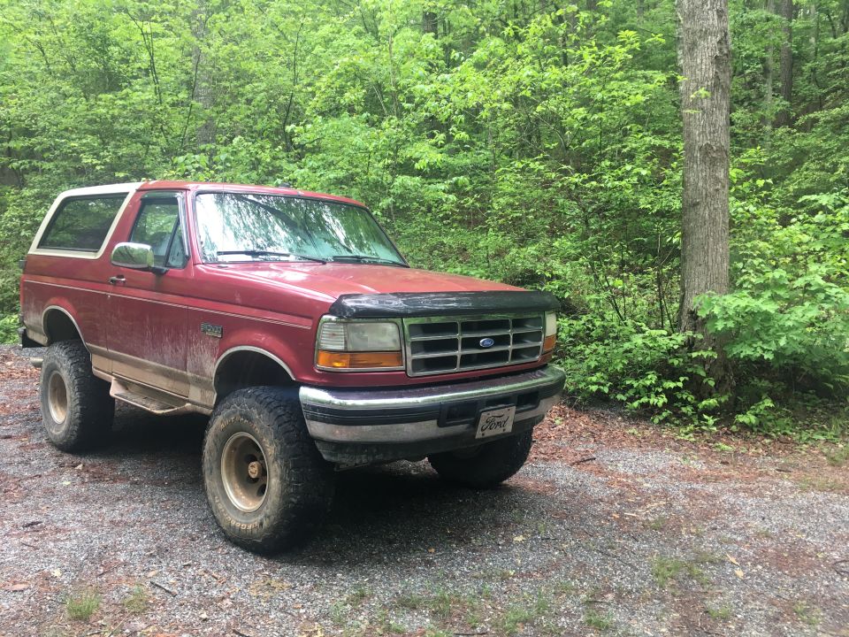 Red 1990s Ford Bronco with white top and large tires parked in a forest.