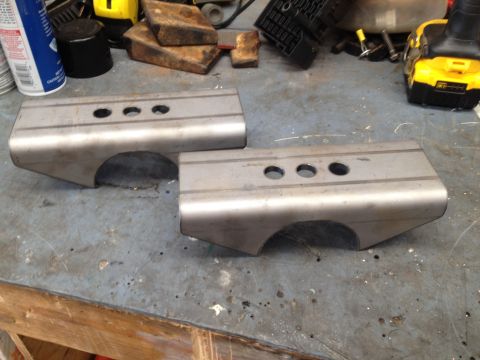 1979 Ford Bronco - Two custom-fabricated steel suspension components with three holes drilled along the top are laid out on a workbench. The components appear…