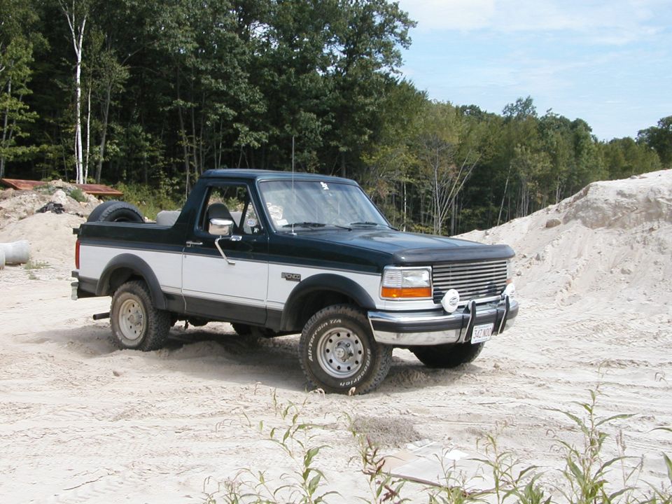Two-tone dark green and white Ford Bronco with a spare tire on the back, parked on sandy terrain