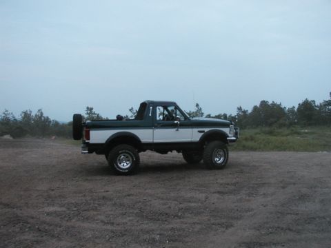 1994 Ford Bronco - This lifted Ford Bronco is shown from the side on a gravelly surface with trees in the background.