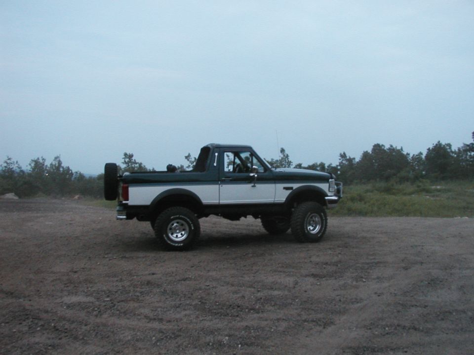 Lifted green and white full-size Bronco, top removed, roll bar and spare tire, side view on gravel