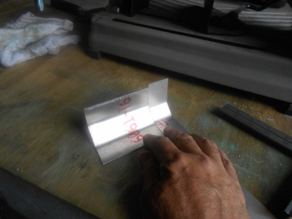 Metal bracket with red markings '91-T909' held by a hand over a wooden surface near a cutting tool