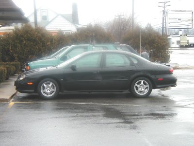 1997 Ford Taurus - A black sedan is parked on a wet asphalt lot next to a green truck on an overcast day.