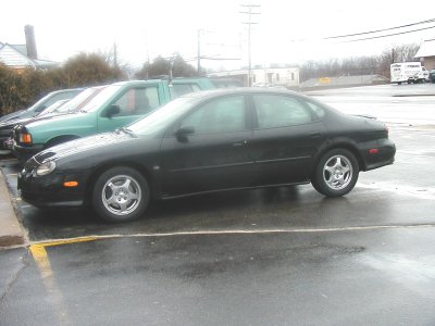 1997 Ford Taurus - A black sedan is parked in a lot next to a green truck.