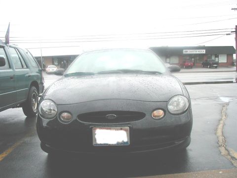 1997 Ford Taurus - A dark 1997 Ford Taurus is parked outside a medical building on a rainy day, with a green SUV visible to its left.