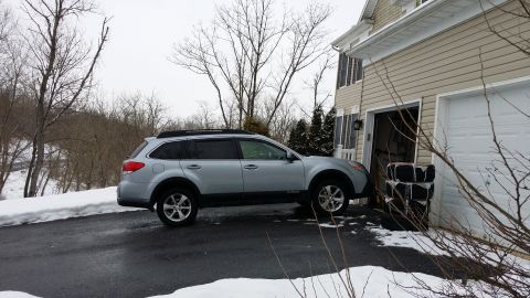 2014 Subaru Outback - Silver Subaru Outback parked on a wet driveway next to a beige house with an open garage door.