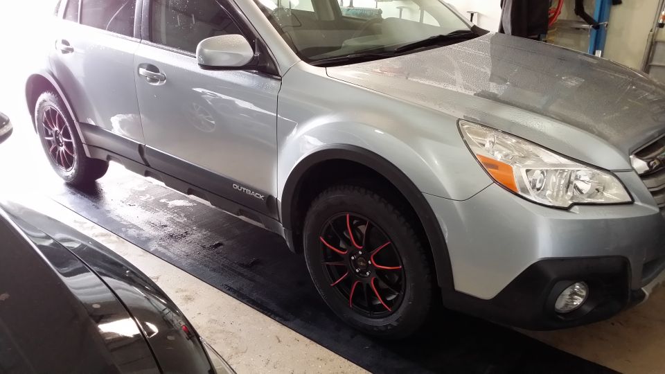 Silver Subaru Outback with black and red wheels, 'Outback' badge visible on lower door trim. Another dark vehicle is partially visible on the left