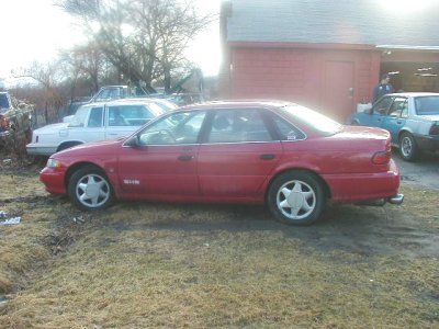 1997 Ford Taurus - Side view of a red 1997 Ford Taurus sedan parked outdoors near a red building.