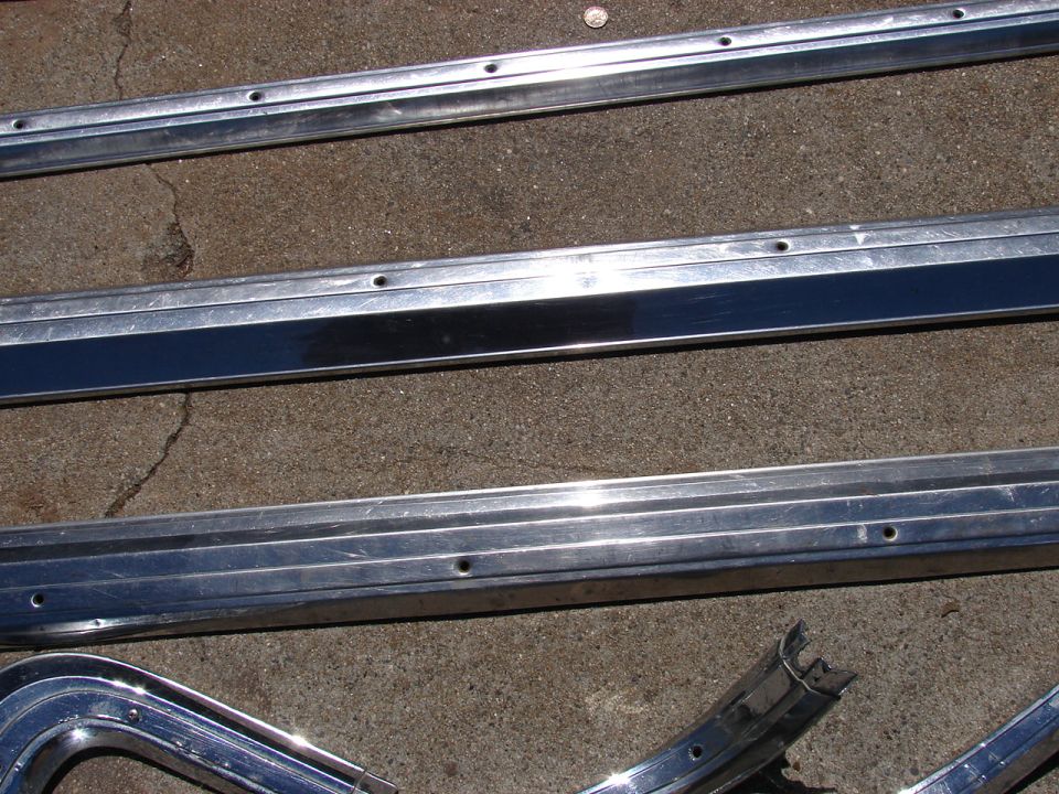 Several long, flat chrome trim pieces with screw holes are laid out on a concrete surface. One piece has a black insert