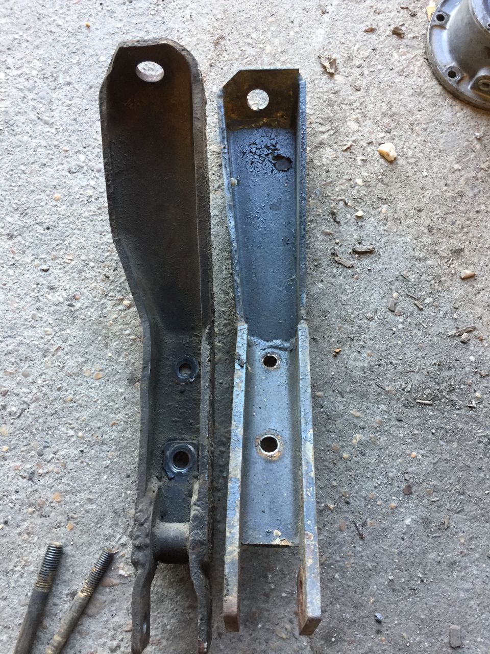 Two metal suspension brackets, one dark and worn, the other lighter and recently painted, lie on a concrete surface