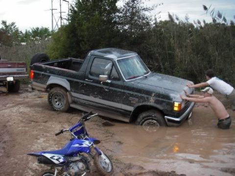 1989 Ford Bronco - A Ford Bronco is stuck in the mud with two people attempting to push it out, while another vehicle waits behind.