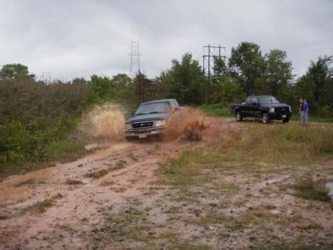 1989 Ford Bronco - A dark SUV drives through a muddy track, kicking up a large splash, while a black pickup truck waits in the background.