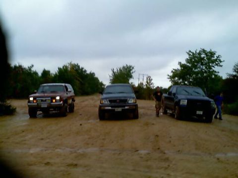 1989 Ford Bronco - Three SUVs and trucks parked on a muddy dirt road with trees in the background.