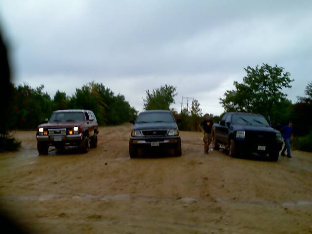 Three SUVs and trucks on a dirt road under a cloudy sky. The leftmost vehicle is a red SUV, possibly a 1997 Ford Bronco. A dark SUV is in the middle, and a dark pickup truck is on the right
