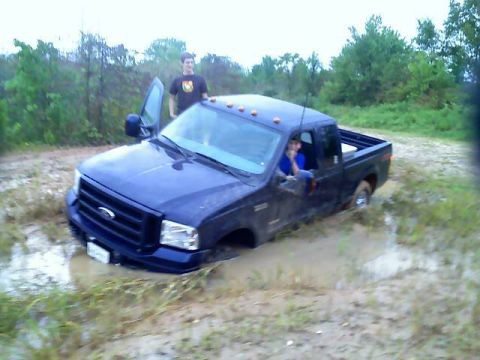 1989 Ford Bronco - A blue pickup truck is stuck deep in mud with two people nearby.