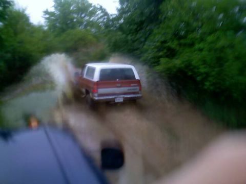 1989 Ford Bronco - 1989 Ford Bronco splashes through water on a dirt road, with another vehicle in the foreground.