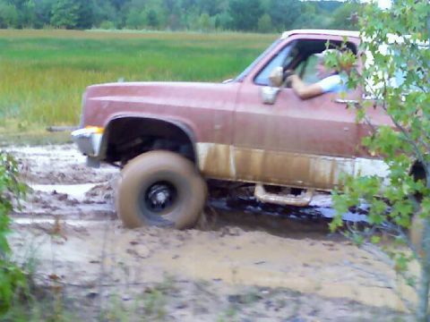 1989 Ford Bronco - A red pickup truck navigates through a muddy field, with its tires deep in the mire.
