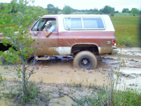 1989 Ford Bronco - A red and white SUV is stuck deep in mud, with its driver visible behind the wheel.
