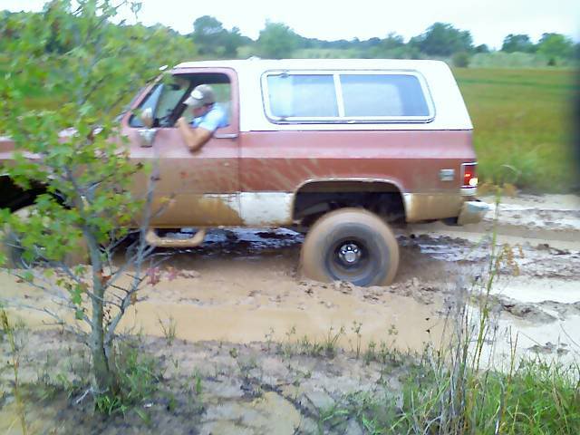 Red and white SUV with a white camper shell stuck in thick mud, driver visible