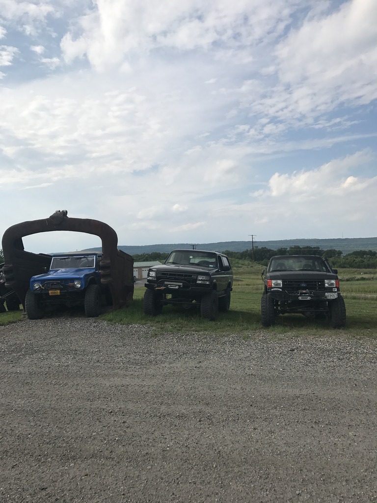 Three SUVs, including a blue 1989 Ford Bronco, parked in front of a large metal sculpture