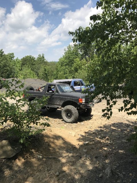 1989 Ford Bronco - A modified black SUV with oversized tires is parked off-road, with another vehicle visible behind it.