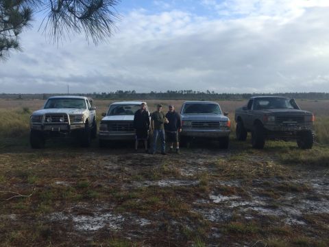 1989 Ford Bronco - A group of friends and their trucks pose for a photo in a grassy, open field under a cloudy sky.