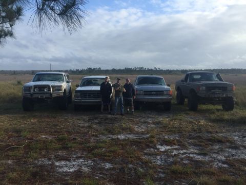 1989 Ford Bronco - Four trucks are parked in a grassy field under a cloudy sky with three men standing in front of the middle two vehicles.