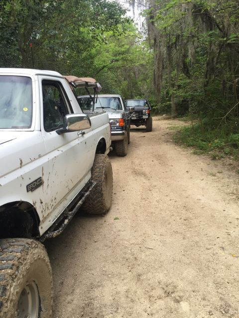 1989 Ford Bronco - A line of SUVs, led by a white Ford Bronco, drives down a dirt path surrounded by trees.