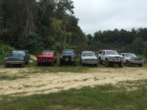1989 Ford Bronco - A lineup of SUVs, including a green 1989 Ford Bronco, parked outdoors on a cloudy day.