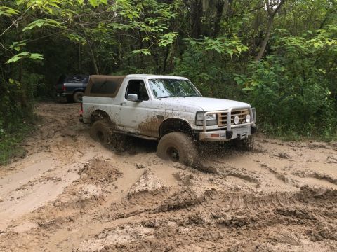 1989 Ford Bronco - A white Ford Bronco navigates a challenging muddy path in a wooded area, with another vehicle following behind.