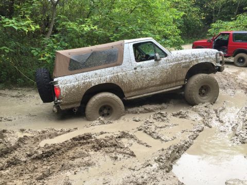 1989 Ford Bronco - Two Ford Broncos navigate a muddy off-road trail, with the white Bronco partially submerged in thick mud.