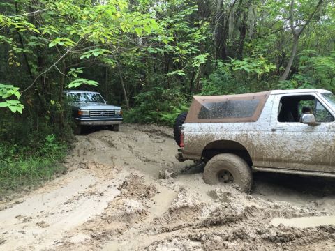 1989 Ford Bronco - Two Ford Broncos navigate a muddy trail, one deeply stuck in the mire.