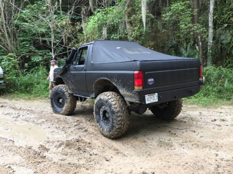 1989 Ford Bronco - This modified Ford Bronco is shown off-road on a muddy path, with a second vehicle partially visible in the background.
