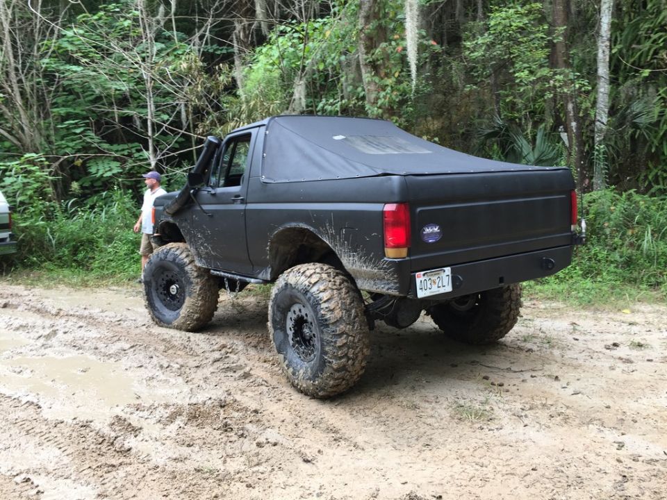 Black Ford Bronco with soft top and big tires on a muddy trail. Another vehicle is partially visible to the left