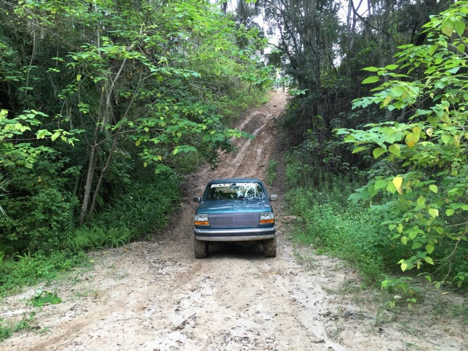 Teal SUV with a chrome grille and a sticker on the windshield drives up a muddy, tree-lined road