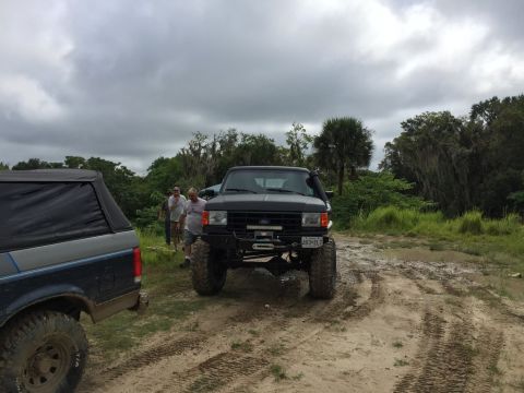 1989 Ford Bronco - Two off-road vehicles are parked on a dirt path with trees and cloudy skies in the background.