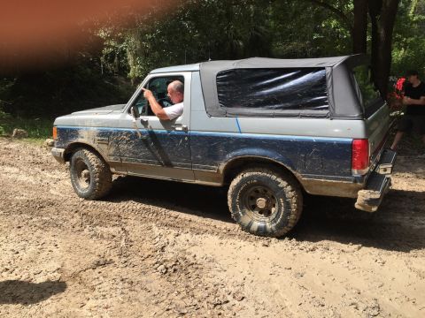 1989 Ford Bronco - Driver points ahead in a muddy 1989 Ford Bronco with a soft top during an off-road excursion.