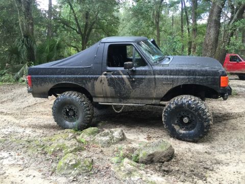 1989 Ford Bronco - A black Ford Bronco navigates a muddy off-road course with a red truck visible in the background.