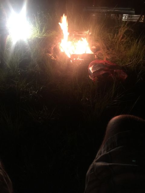 1989 Ford Bronco - A bonfire burns brightly at night, illuminating the surrounding grass and a person's leg in the foreground.