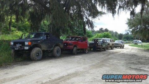 1989 Ford Bronco - Multiple off-road vehicles are parked in a line on a dirt path, with trees overhead.